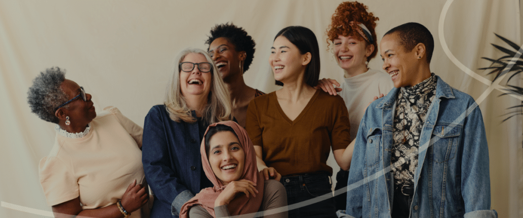 Group of six diverse women standing beside each other and smiling against a cream coloured backdrop.